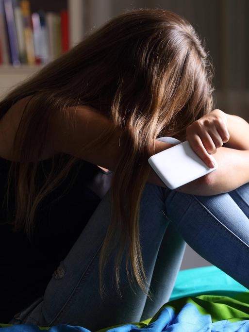 Ein Mädchen sitzt mit dem Smartphone in der Hand traurig auf dem Bett.