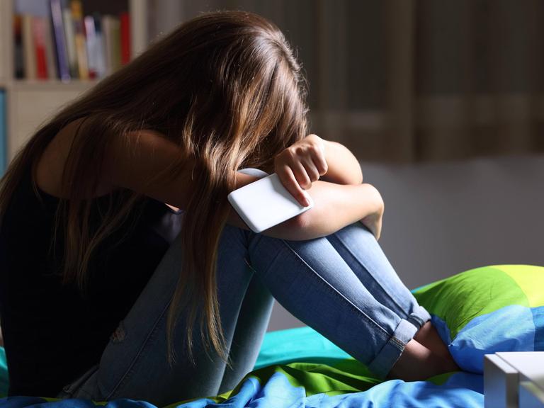 Ein Mädchen sitzt mit dem Smartphone in der Hand traurig auf dem Bett.