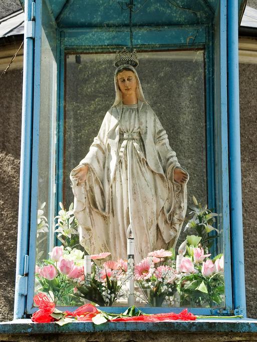 Die Madonnenstatue zeigt Maria in einem weißen Kleid und mit Krone. Sie hat die Arme ausgebreitet.