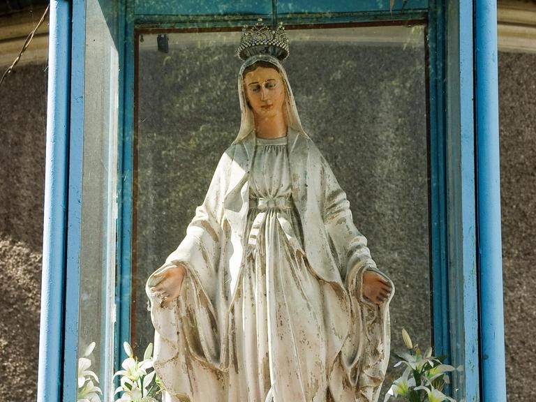 Die Madonnenstatue zeigt Maria in einem weißen Kleid und mit Krone. Sie hat die Arme ausgebreitet.