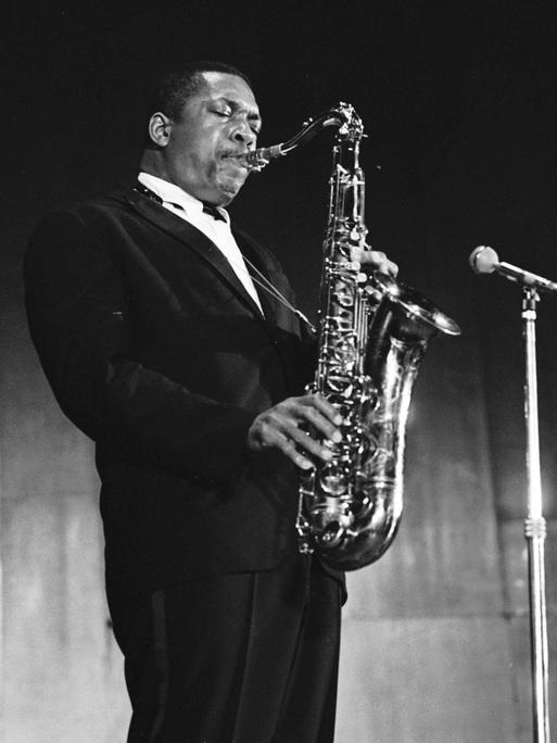 John Coltrane steht im schwarzen Anzug auf der Bühne und spielt Saxofon vor einem Mikrofon. John Coltrane steht im schwarzen Anzug auf der Bühne und spielt Saxofon vor einem Mikrofon.