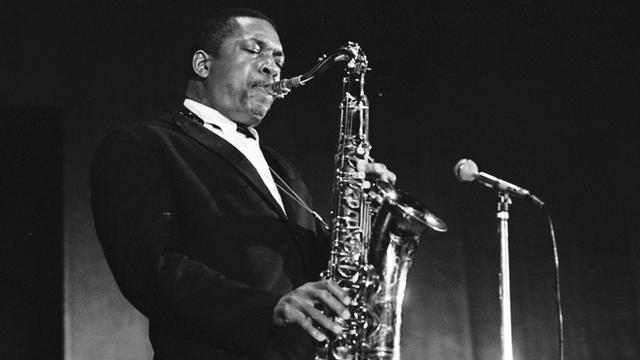 John Coltrane steht im schwarzen Anzug auf der Bühne und spielt Saxofon vor einem Mikrofon. John Coltrane steht im schwarzen Anzug auf der Bühne und spielt Saxofon vor einem Mikrofon.