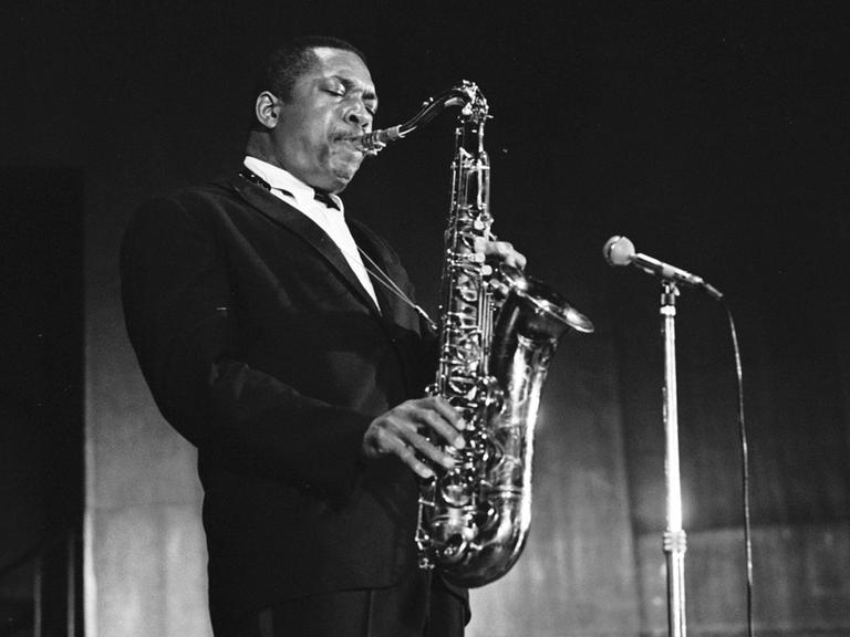John Coltrane steht im schwarzen Anzug auf der Bühne und spielt Saxofon vor einem Mikrofon. John Coltrane steht im schwarzen Anzug auf der Bühne und spielt Saxofon vor einem Mikrofon.