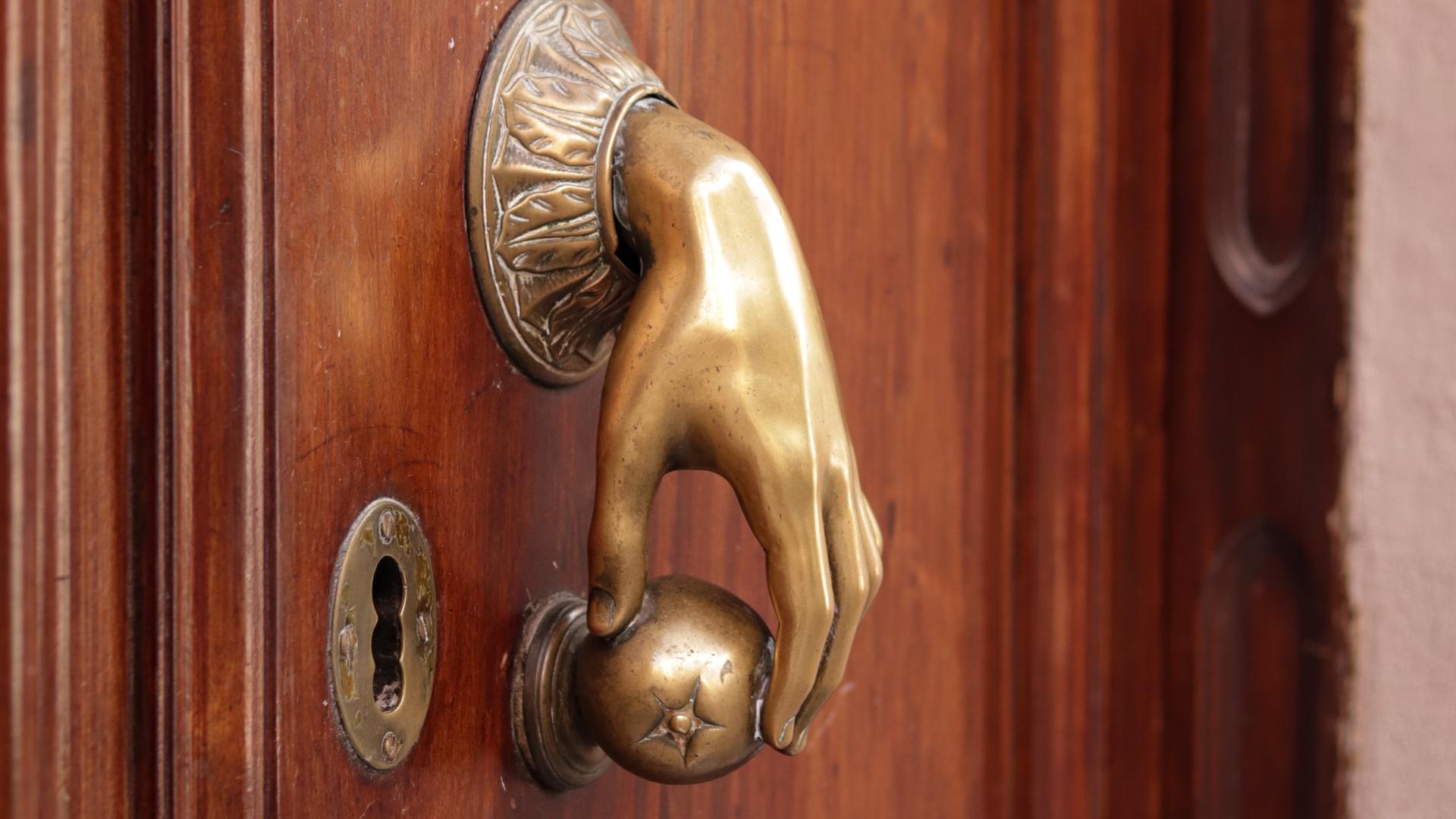 Ein besonderer Türgriff: eine Hand aus Messing greift eine kleine runde Kugel. Mit diesem ungewöhnlichen Griff lässt sich eine braune Holztür öffnen.