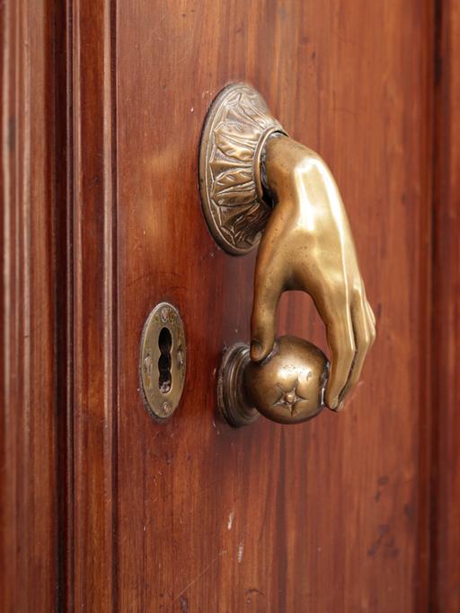 Ein besonderer Türgriff: eine Hand aus Messing greift eine kleine runde Kugel. Mit diesem ungewöhnlichen Griff lässt sich eine braune Holztür öffnen.