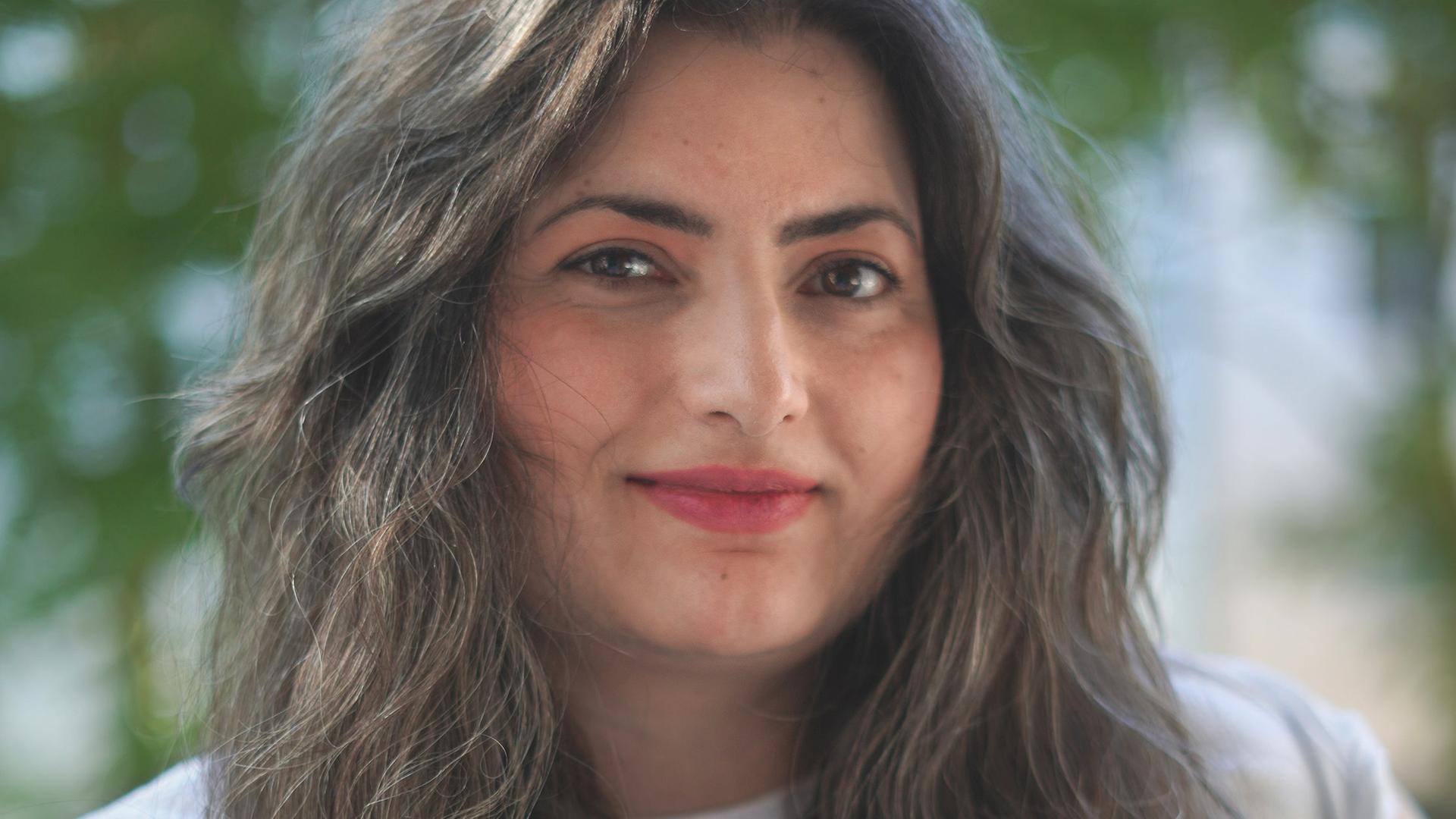 Porträt von Şehnaz Dost. Sie hat langes dunkelbraunes Haar und trägt ein weißes T-Shirt.                               