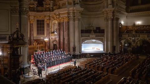 Ein Blick in eine große, hohe Kirche, den Berliner Dom. Im Altarraum steht ein Chor, alle schwarz gekleidet. Links daneben einige Blechbläser. Vor dem Chor steht der Chorleiter, er hält die Arme erhöht, gibt Zeichen. Die Kirchenbänke sind gefüllt mit Zuschauern.