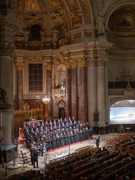 Ein Blick in eine große, hohe Kirche, den Berliner Dom. Im Altarraum steht ein Chor, alle schwarz gekleidet. Links daneben einige Blechbläser. Vor dem Chor steht der Chorleiter, er hält die Arme erhöht, gibt Zeichen. Die Kirchenbänke sind gefüllt mit Zuschauern.