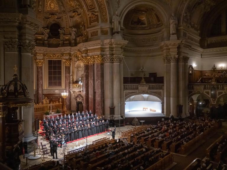 Ein Blick in eine große, hohe Kirche, den Berliner Dom. Im Altarraum steht ein Chor, alle schwarz gekleidet. Links daneben einige Blechbläser. Vor dem Chor steht der Chorleiter, er hält die Arme erhöht, gibt Zeichen. Die Kirchenbänke sind gefüllt mit Zuschauern.