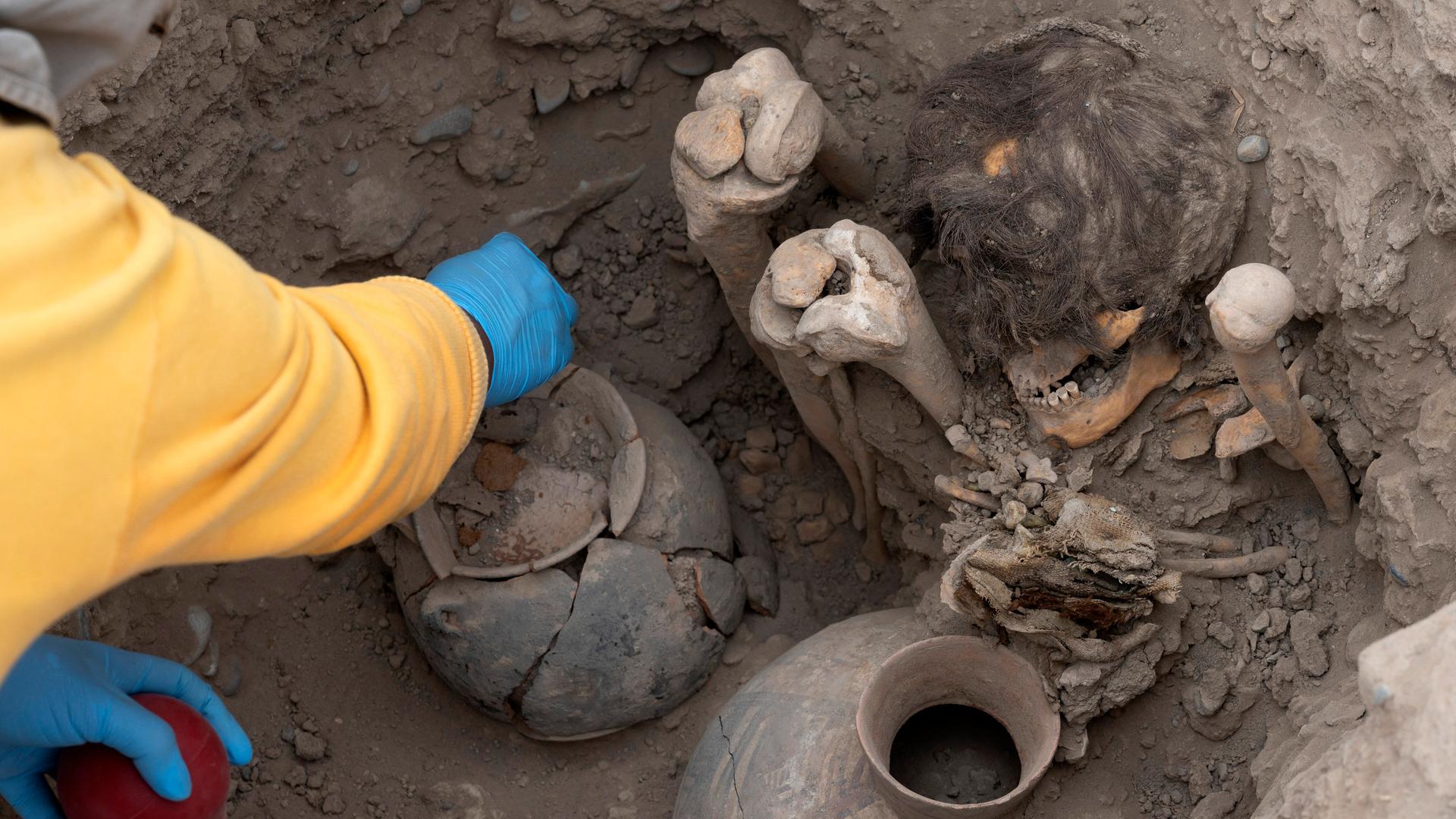 Ein Person befreit die archäologischen Funde von Erde und Staub. Man sieht eine Mumie mit angewinkelten Beinen und intaktem schwarzen Haar.