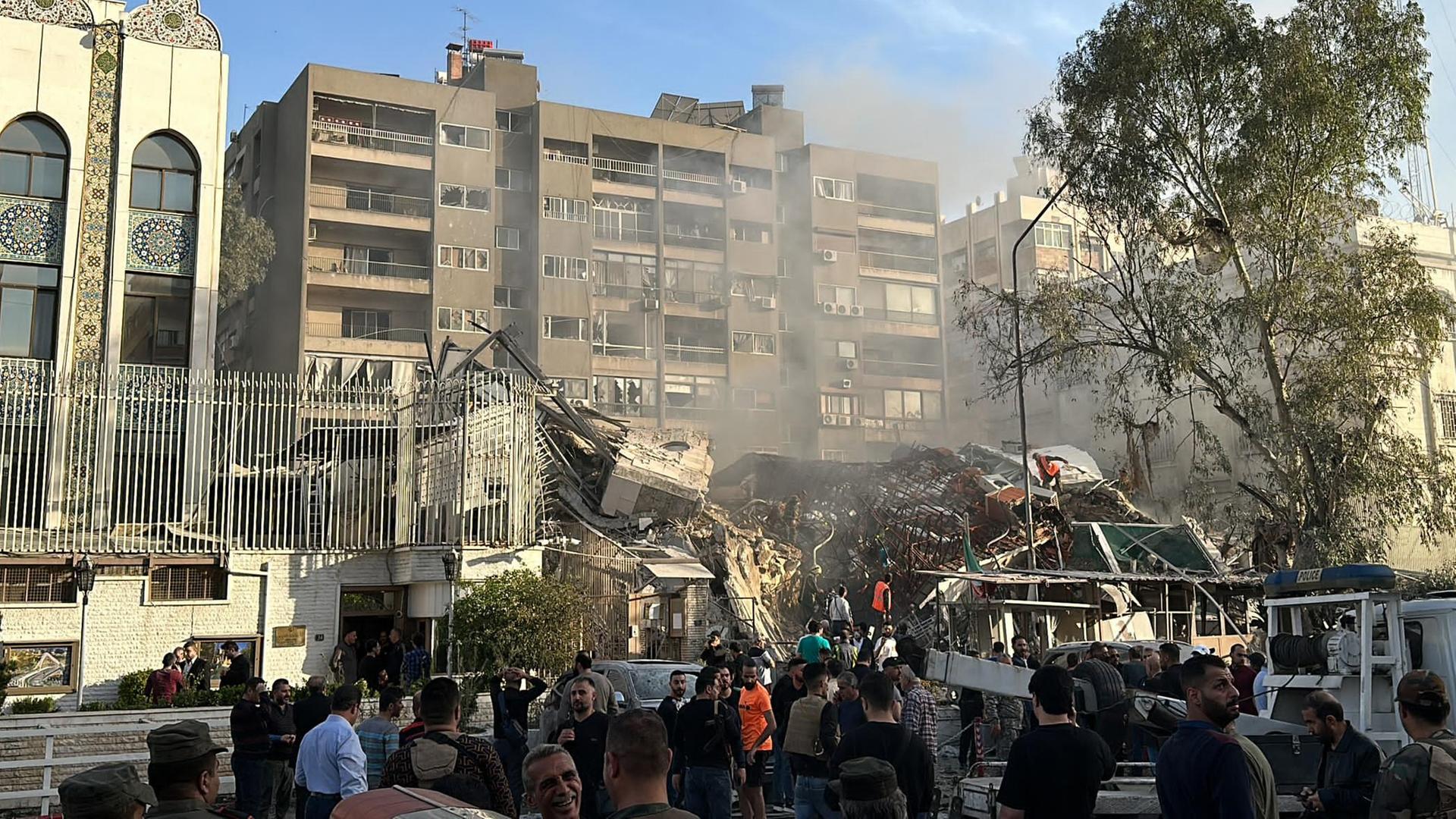 Menschen stehen vor einem komplett eingestürzten Gebäude, Retter versuchen, die Trümmer abzusuchen.