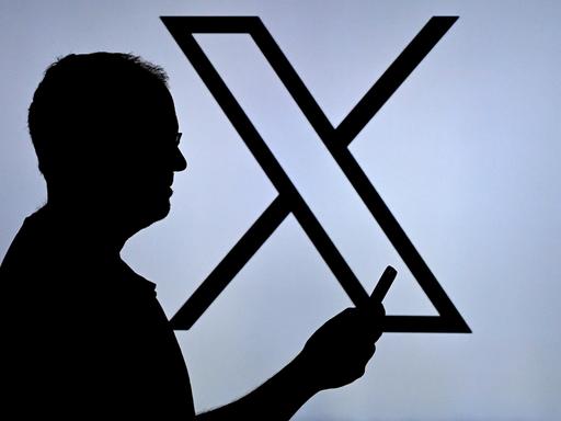 Ein Mann blickt auf sein Handy, im Hintergrund ist ein großes X zu sehen. Ein Mann blickt auf sein Handy, im Hintergrund ist ein großes X zu sehen.
