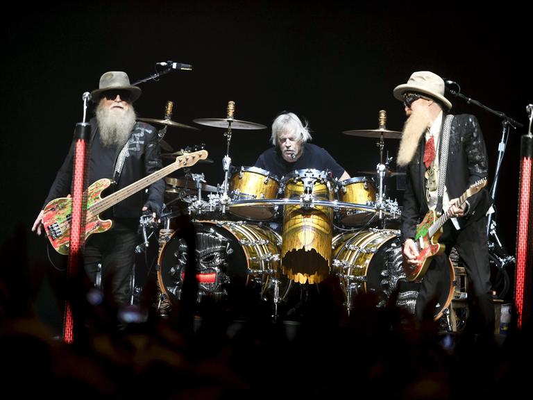 Die Band ZZ Top bei einem Auftritt in Helsinki: Rechts und links ein Mann mit langem Bart und einer E-Gitarre. In der Mitte sitzt ein dritter Mann an einem Schlagzeug, ohne Bart. Die Band ZZ Top bei einem Auftritt in Helsinki: Rechts und links ein Mann mit langem Bart und einer E-Gitarre. In der Mitte sitzt ein dritter Mann an einem Schlagzeug, ohne Bart.