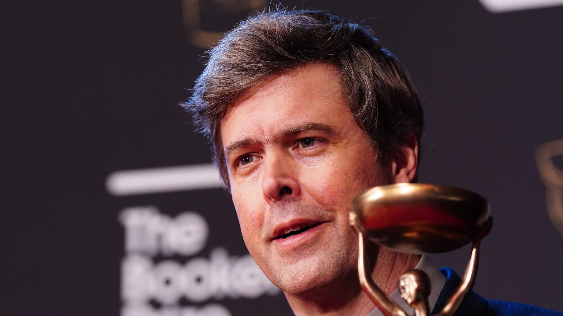 Schriftsteller David Szalay hält die Statue des Booker Prize lächelnd in die Kamera. 