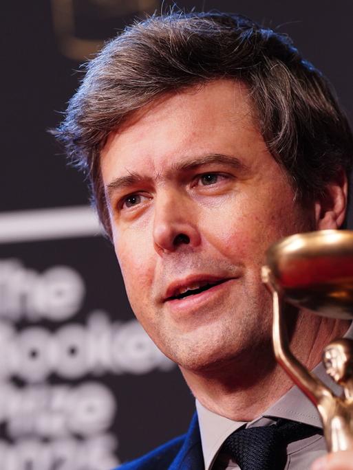 Schriftsteller David Szalay hält die Statue des Booker Prize lächelnd in die Kamera. 