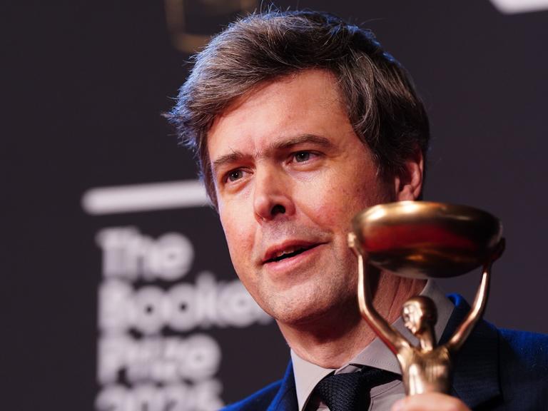 Schriftsteller David Szalay hält die Statue des Booker Prize lächelnd in die Kamera. 