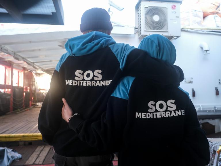 Ein Mann und eine Frau stehen Arm in Arm auf einem Schiff und tragen Anoraks mit der Aufschrift SOS Mediterranee