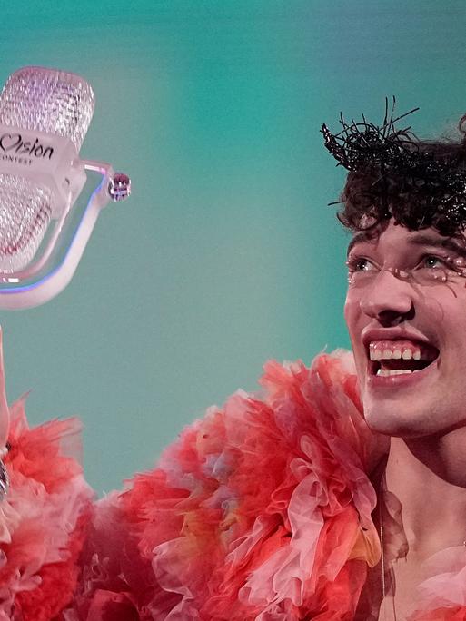 Eine Person hält ein Mikrofon aus Glas in die Höhe. Die Person trägt pink und orangefarbene Rüschen, hat einen dunklen, lockigen Kurzhaarschnitt und lacht übers ganze Gesicht.