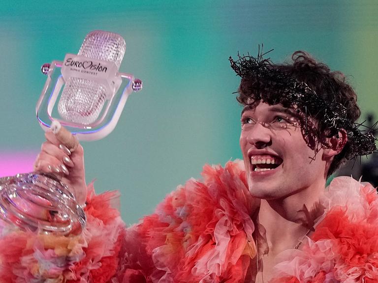 Eine Person hält ein Mikrofon aus Glas in die Höhe. Die Person trägt pink und orangefarbene Rüschen, hat einen dunklen, lockigen Kurzhaarschnitt und lacht übers ganze Gesicht.