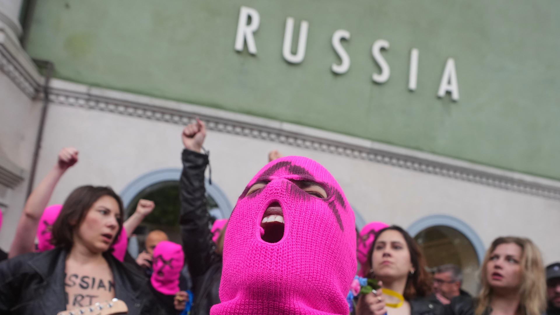 Eine Person mit rosa Strickmaske schreit. Hinter ihr sind barbusige Frauen mit Lederjacken. An einer Hauswand steht der Schriftzug "Russia"