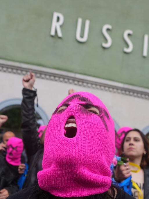 Eine Person mit rosa Strickmaske schreit. Hinter ihr sind barbusige Frauen mit Lederjacken. An einer Hauswand steht der Schriftzug "Russia"