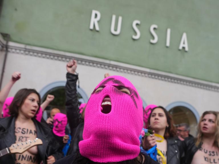 Eine Person mit rosa Strickmaske schreit. Hinter ihr sind barbusige Frauen mit Lederjacken. An einer Hauswand steht der Schriftzug "Russia"