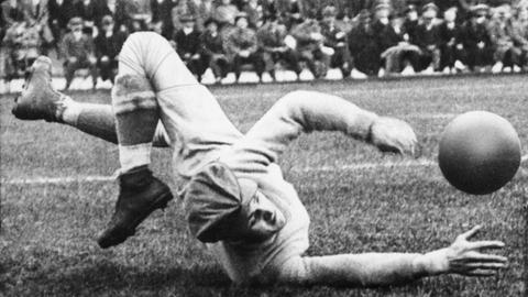 Schwarzweißfoto von einem historischen Fußballspiel. Ein Torhüter liegt auf dem Fußballfeld und fängt einen Fußball. 