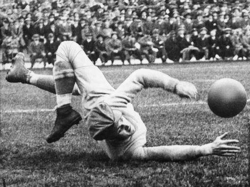 Schwarzweißfoto von einem historischen Fußballspiel. Ein Torhüter liegt auf dem Fußballfeld und fängt einen Fußball. 