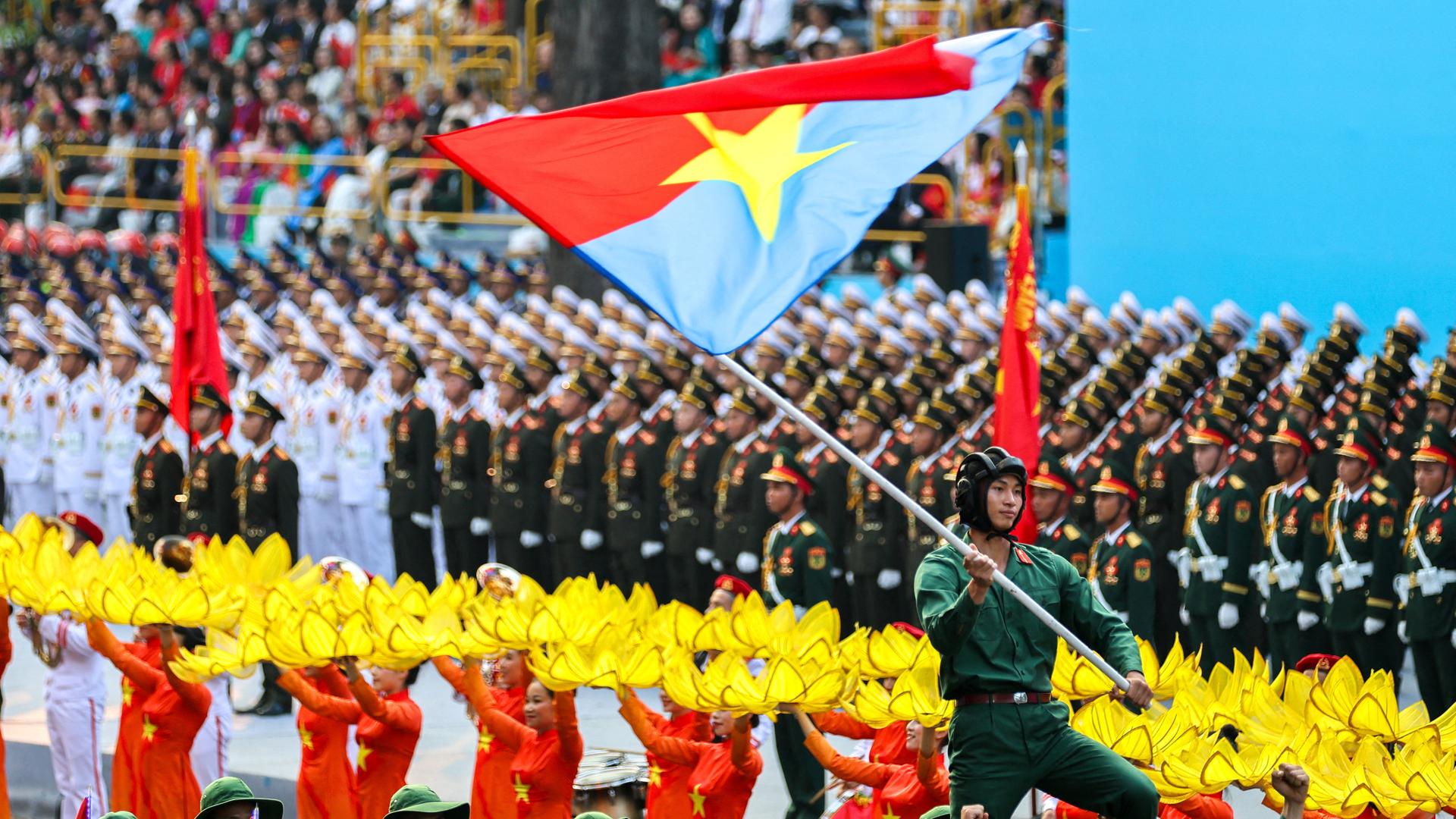 Das Bild zeigt eine farbenfrohe Militärparade in Ho-Chi-Minh-Stadt. Ein Soldat schwenkt eine rot-blaue Flagge mit einem gelben Stern.
