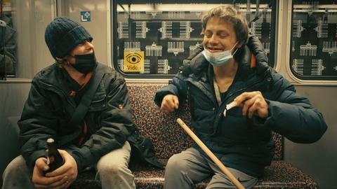 Im Still aus "Hausnummer Null" sitzen zwei Männer mit Bierflaschen nebeneinander in der U-Bahn und lachen miteinander.
