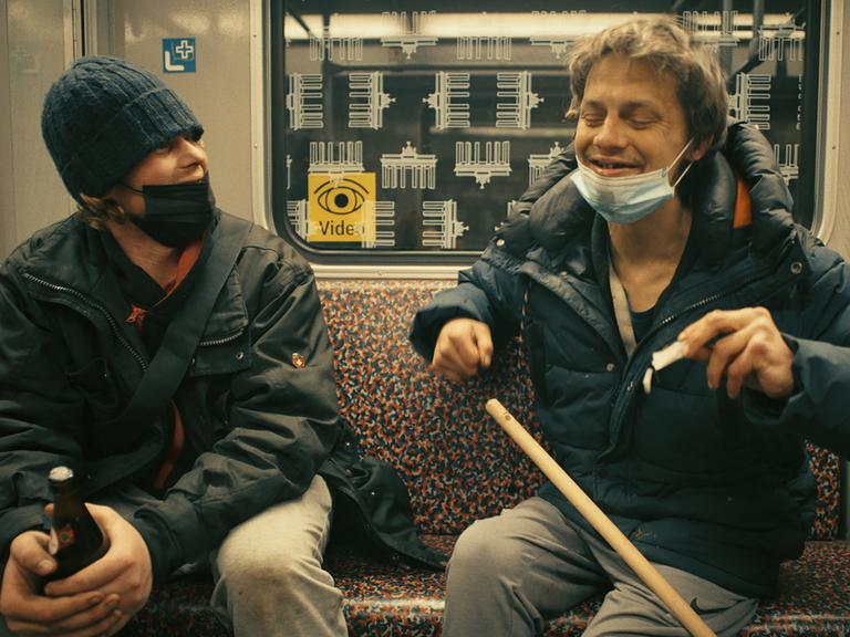 Im Still aus "Hausnummer Null" sitzen zwei Männer mit Bierflaschen nebeneinander in der U-Bahn und lachen miteinander.