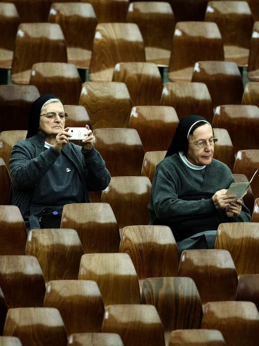 Eine Nonne fotografiert mit ihrem Smartphone während der Feierlichkeiten zum Jubiläum der Errichtung der Bischofssynode im Vatikan. Schräg vor ihr sitzt eine zweite Nonne, die in einem Flyer blättert. (Symbolbild) Eine Nonne fotografiert mit ihrem Smartphone während der Feierlichkeiten zum Jubiläum der Errichtung der Bischofssynode im Vatikan. Schräg vor ihr sitzt eine zweite Nonne, die in einem Flyer blättert. (Symbolbild)