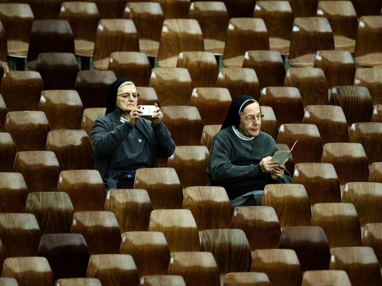Eine Nonne fotografiert mit ihrem Smartphone während der Feierlichkeiten zum Jubiläum der Errichtung der Bischofssynode im Vatikan. Schräg vor ihr sitzt eine zweite Nonne, die in einem Flyer blättert. (Symbolbild) Eine Nonne fotografiert mit ihrem Smartphone während der Feierlichkeiten zum Jubiläum der Errichtung der Bischofssynode im Vatikan. Schräg vor ihr sitzt eine zweite Nonne, die in einem Flyer blättert. (Symbolbild)