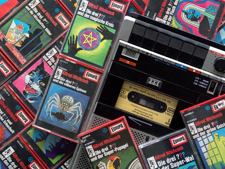 Zahlreiche Kassetten der Hörspielserie "Die drei Fragezeichen" liegen neben einem Kassettenrekorder. 