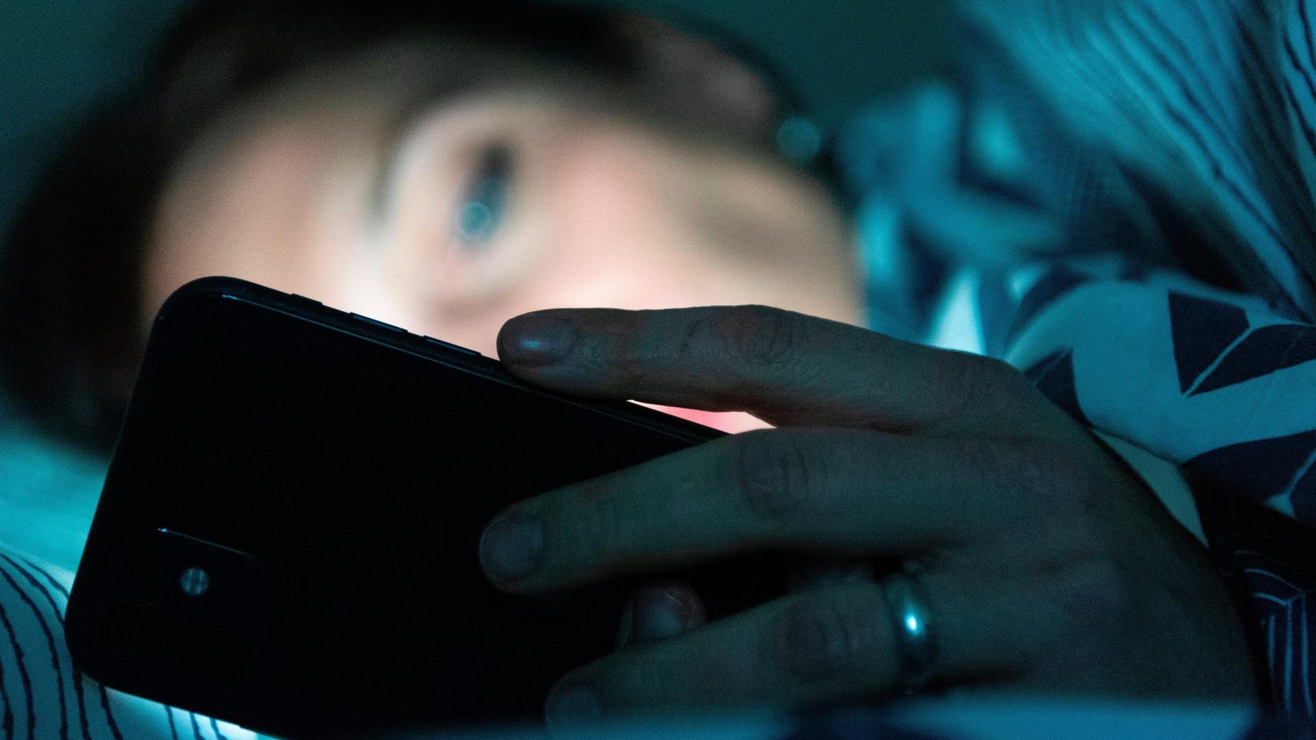 Eine Person liegt im Bett und schaut auf ein Smartphone. 