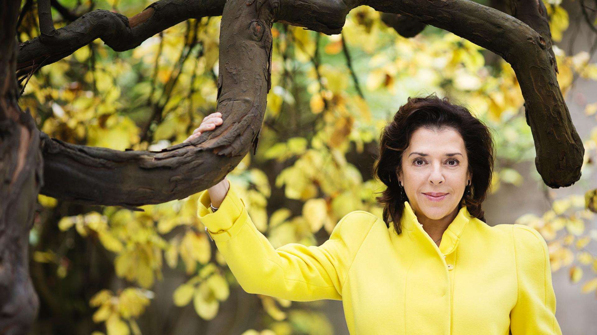 Elena Bashkirova steht in einem gelben Kostüm unter einem gelb belaubten Baum mit markanten, dunklen Ästen.