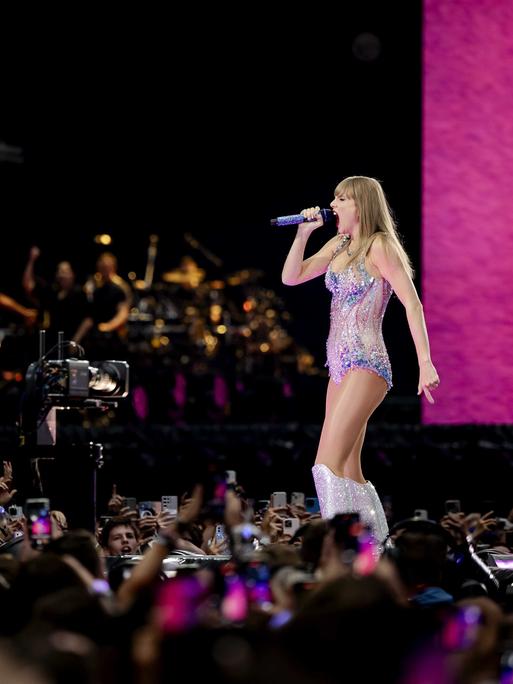 Taylor Swift während ihres Auftritts vor rund 55.000 Menschen in einer Konzerthalle. Sie trägt einen Glitzerbody und Glitzerstiefel, um sie stehen Fans dicht an dicht, manchen filmen mit ihren Handys.