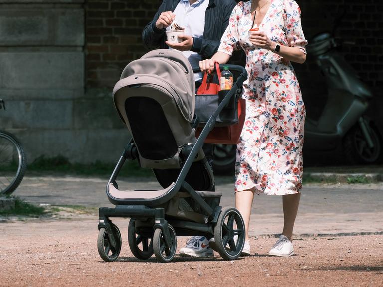 Eine Frau schiebt einen Kinderwagen. Direkt hinter ihr geht ein Mann. 