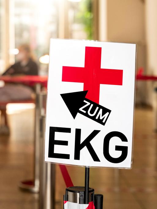 "Zum EKG!" steht auf einem Schild einer Arztpraxis - im Hintergrund zu sehen sind medizinische Fachangestellte. (Symbolbild) 