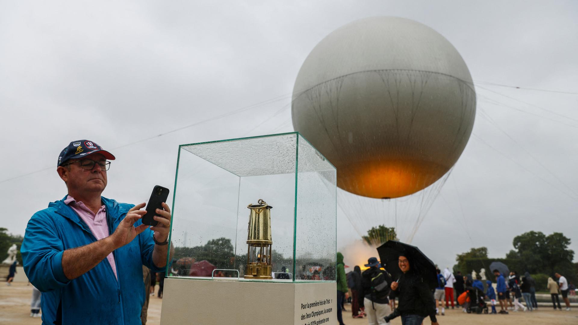 Ein Mann fotografiert mit seinen Handy einen Glaskasten. Im Hintergrund ein Heißluftballon. Ein Mann fotografiert mit seinen Handy einen Glaskasten. Im Hintergrund ein Heißluftballon.