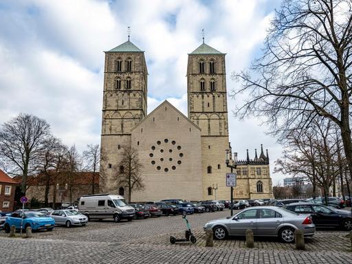 Blick auf das hellbraune Kirchengebäude vor einem großen Platz mit Pflastersteinen, auf dem viele Autos geparkt sind, und auf dem kahle Bäume stehen. Der Himmel darüber ist bewölkt.