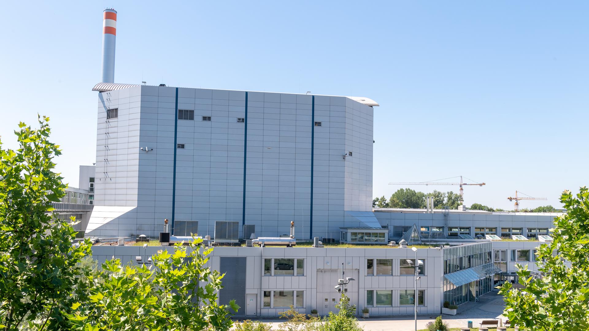 Der Garchinger Forschungsreaktor FRM II auf dem Gelände der Technischen Universität München. Ein weißes gebäude mit einem höheren, fast fensterlosem Gebäude im Hintergrund und Bäumen im Vordergrund. 