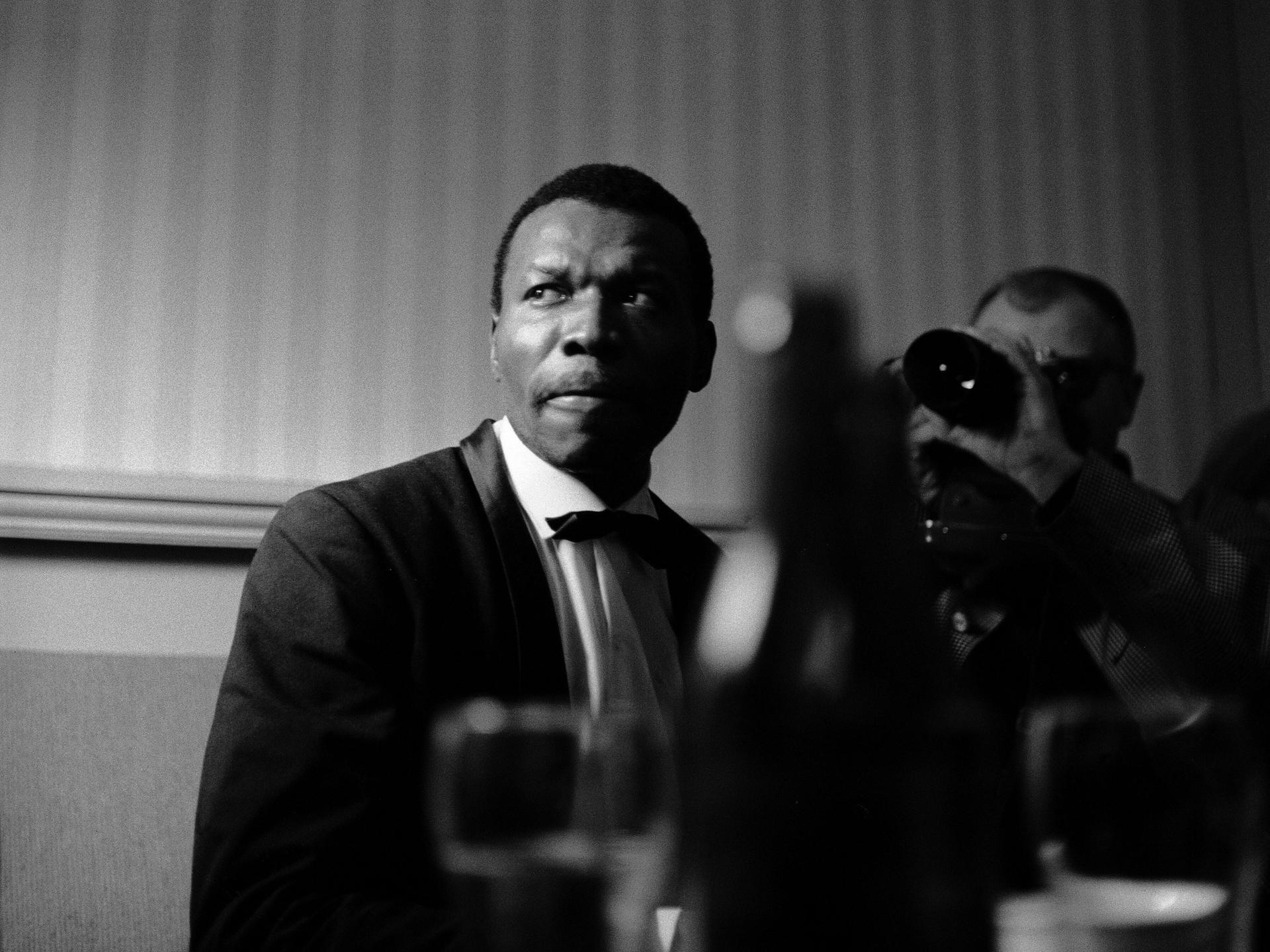 Schwarzweißaufnahme von John Coltrane, der vor einer Wand steht und ernst ins Off blickt. Im Vordergrund ist unscharf ein Musikinstrument zu erkennen. Schwarzweißaufnahme von John Coltrane, der vor einer Wand steht und ernst ins Off blickt. Im Vordergrund ist unscharf ein Musikinstrument zu erkennen.