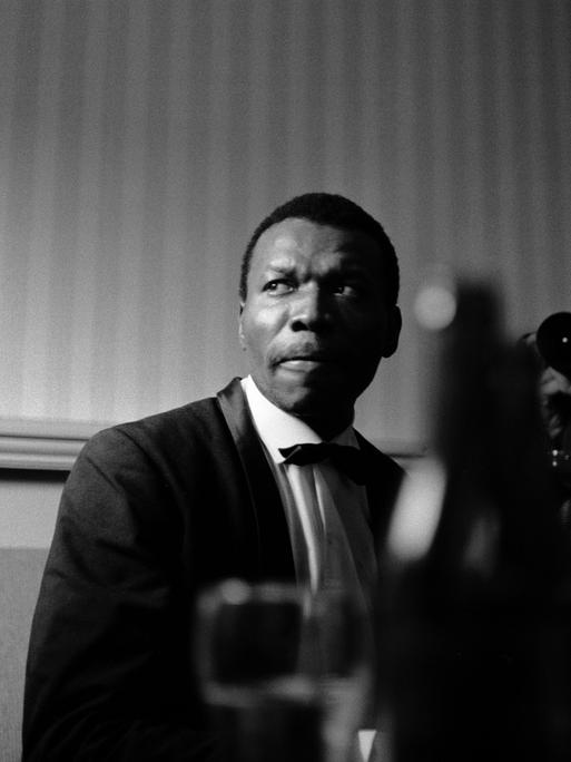 Schwarzweißaufnahme von John Coltrane, der vor einer Wand steht und ernst ins Off blickt. Im Vordergrund ist unscharf ein Musikinstrument zu erkennen.
