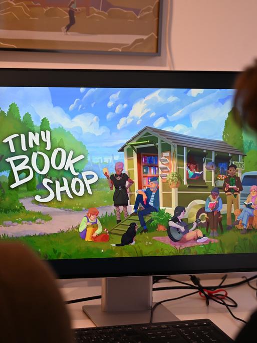 Die Computerspiele-Entwickler David Zapfe-Wildemann (l) und Raven Rusch von dem Kölner Independent-Gamingstudio Neoludic sitzen in ihrem Büro vor einem Bildschirm, auf dem die Startseite des Spiels Tiny Bookshop zu sehen ist. 