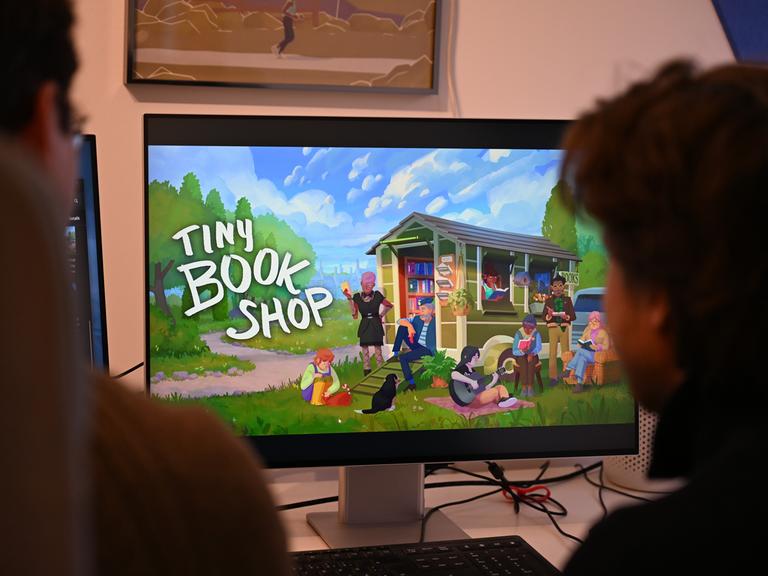 Die Computerspiele-Entwickler David Zapfe-Wildemann (l) und Raven Rusch von dem Kölner Independent-Gamingstudio Neoludic sitzen in ihrem Büro vor einem Bildschirm, auf dem die Startseite des Spiels Tiny Bookshop zu sehen ist. 