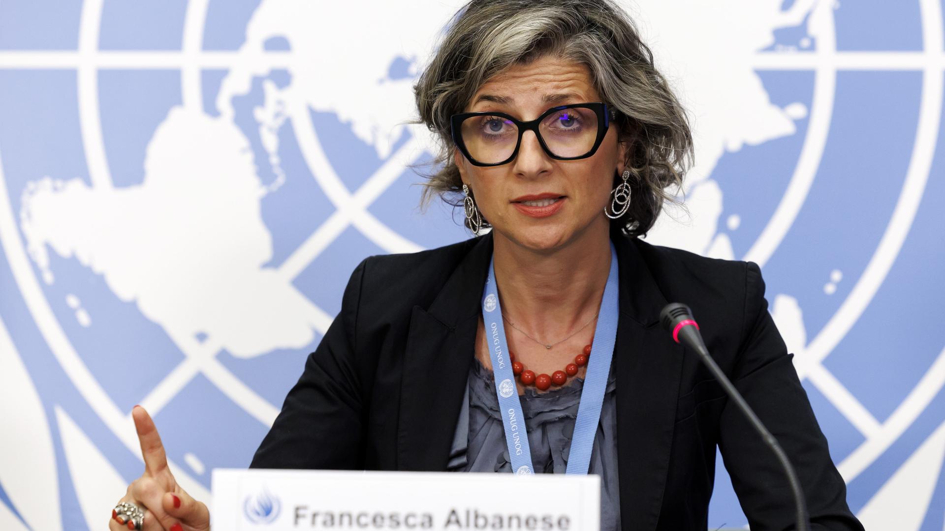 Francesca Albanese sitzt an einem Tisch, spricht in ein Mikrofon und hebt dabei den Zeigefinger der rechten Hand. Im Hintergrund ist eine Weltkarte zu sehen.