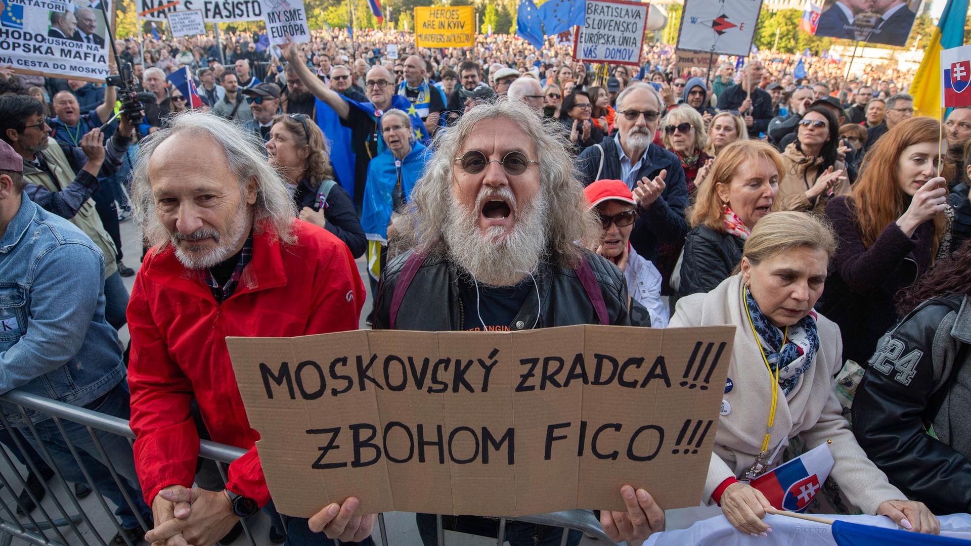 Der bärtige Mann mit dem Plakat steht vorne in der Bildmitte an einem Absperrgitter. Neben und hinter ihm viele weitere Demonstranten. 