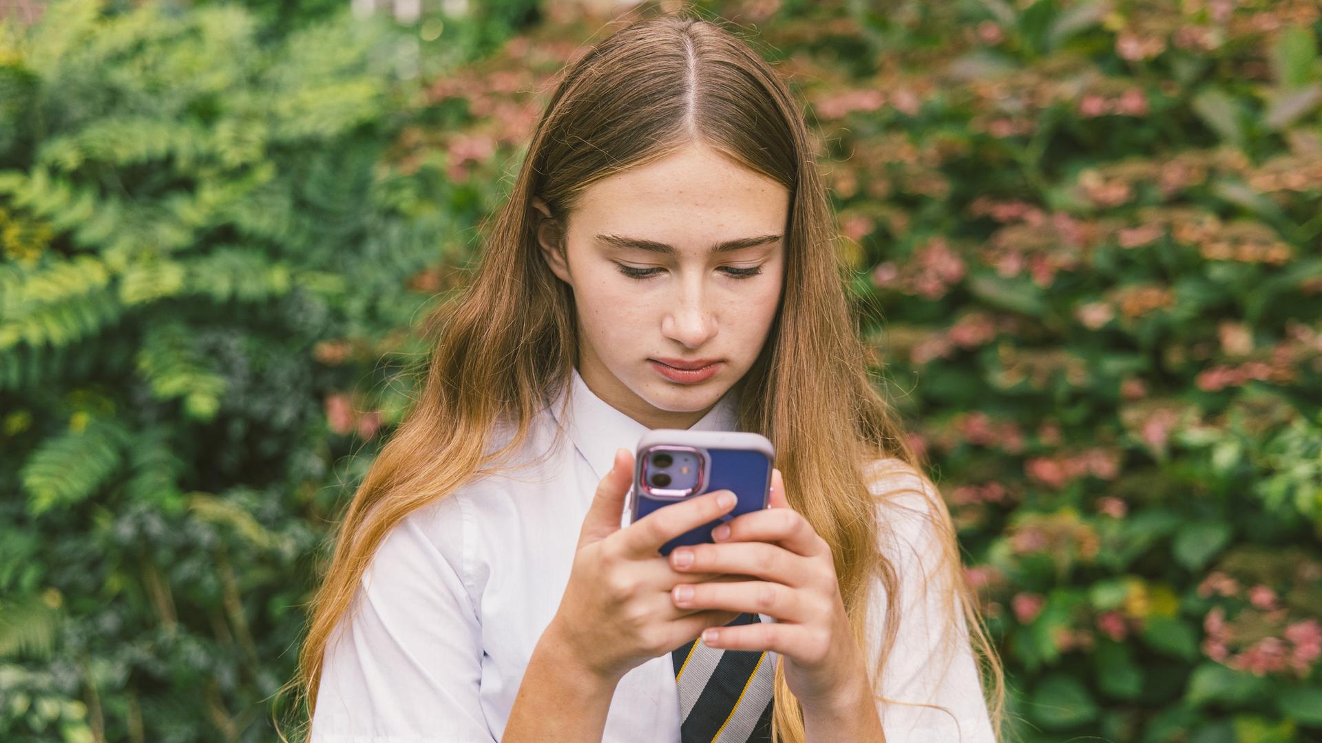 Eine Schülerin in Schuluniform die ernst guckt und auf ihr Smartphone mit bunter Hülle schaut. Eine Schülerin in Schuluniform die ernst guckt und auf ihr Smartphone mit bunter Hülle schaut.