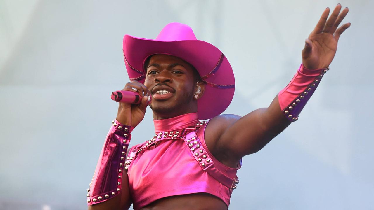 Lil Nas X steht auf einer Bühne. Er trägt einen pinken Cowboy-Hut, ein pinkes Top und ein pinkes Röckchen. In der Hand hält er ein pinkes Mikrofon.